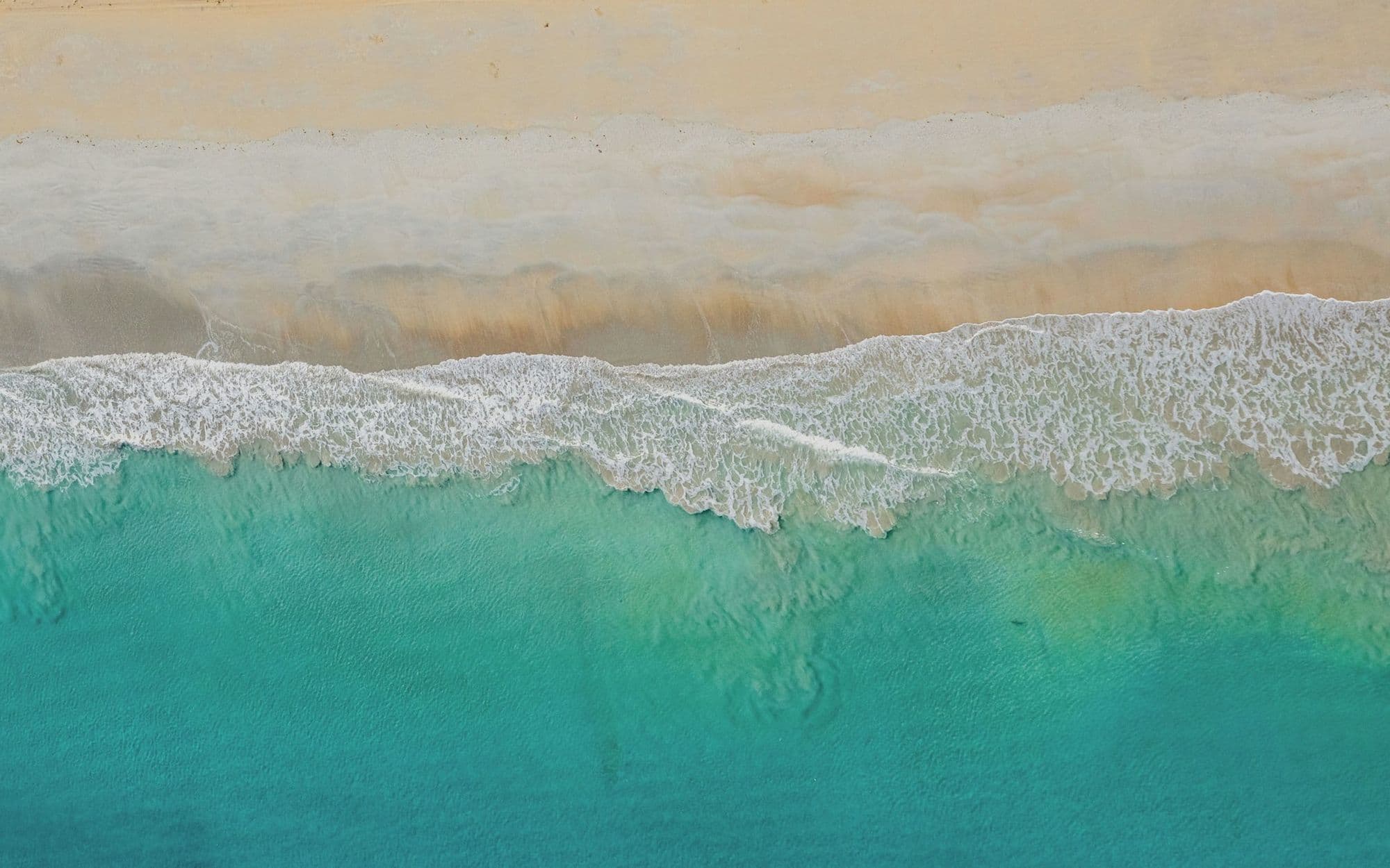 Turquoise waves meeting a sandy shoreline, seen from above