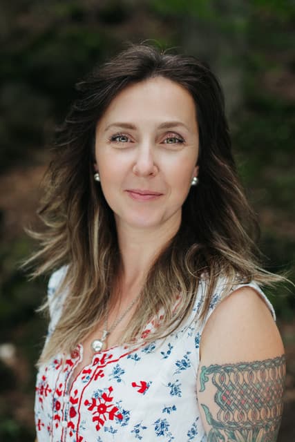 Mariya Garnet in a white floral blouse, looking toward the camera in a forest, Registered Psychotherapist (Qualifying)