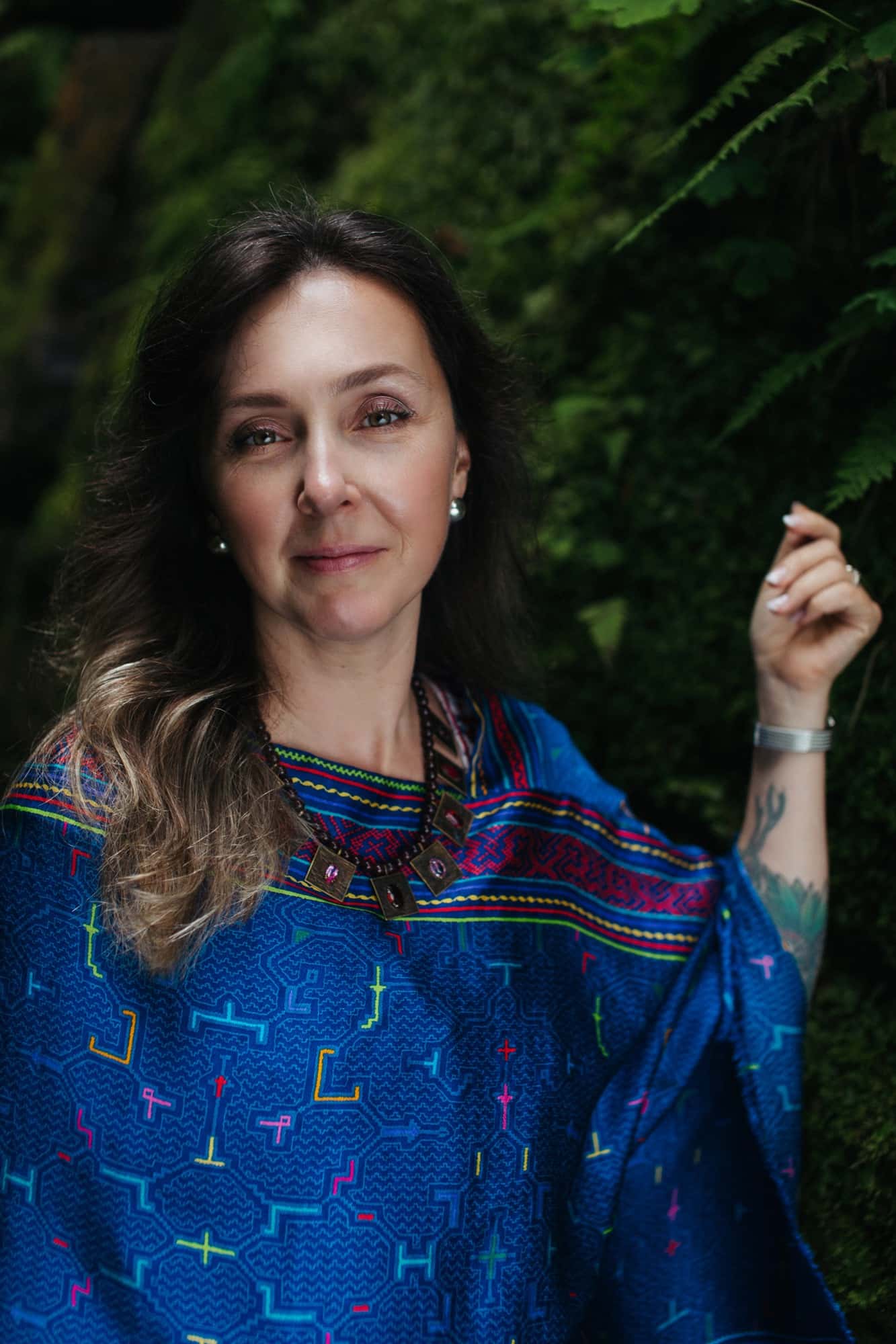 Mariya Garnet smiling and looking toward the camera in a cobalt patterned shawl among ferns