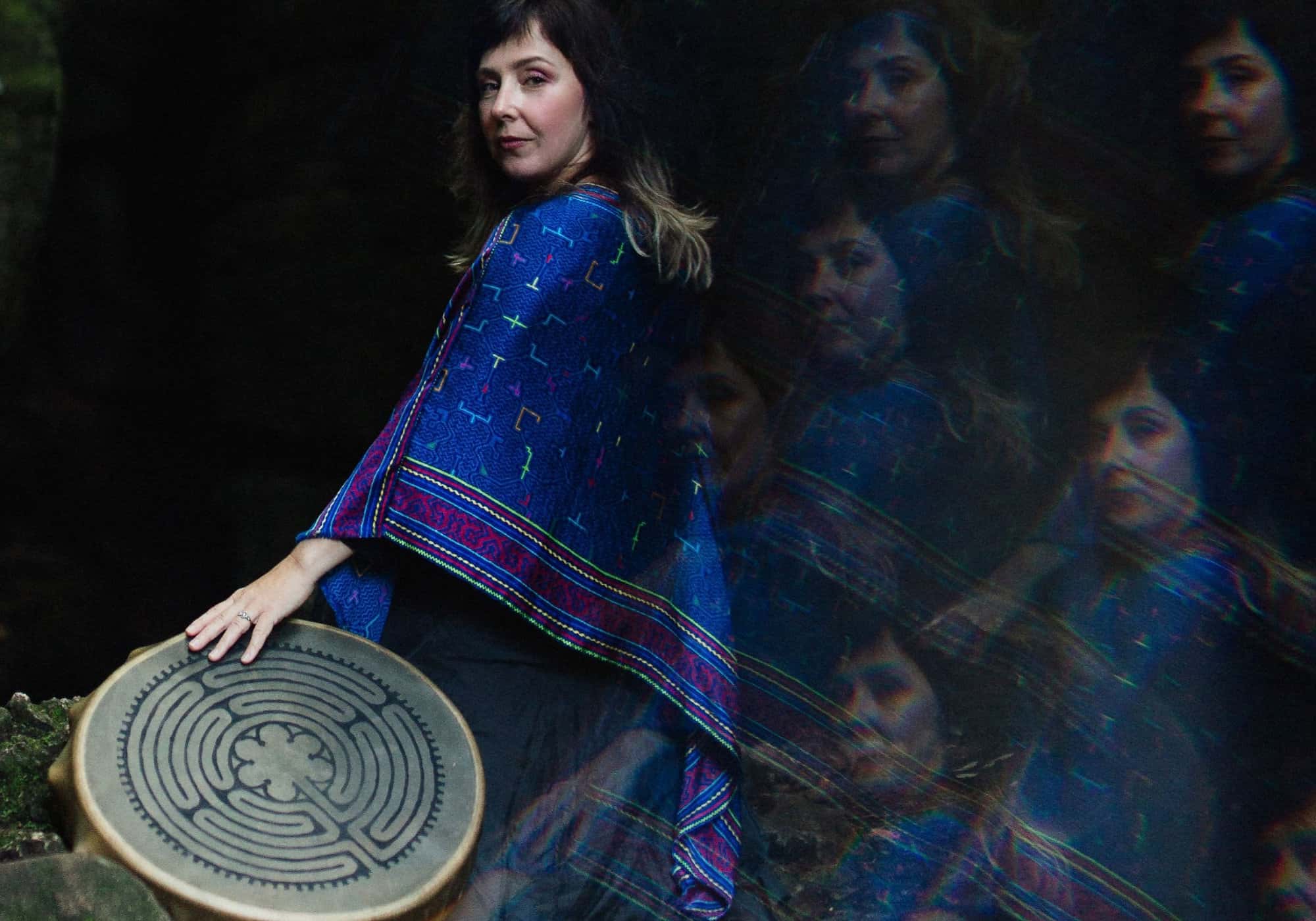 Mariya Garnet in a cobalt shawl holding a labyrinth drum in low light