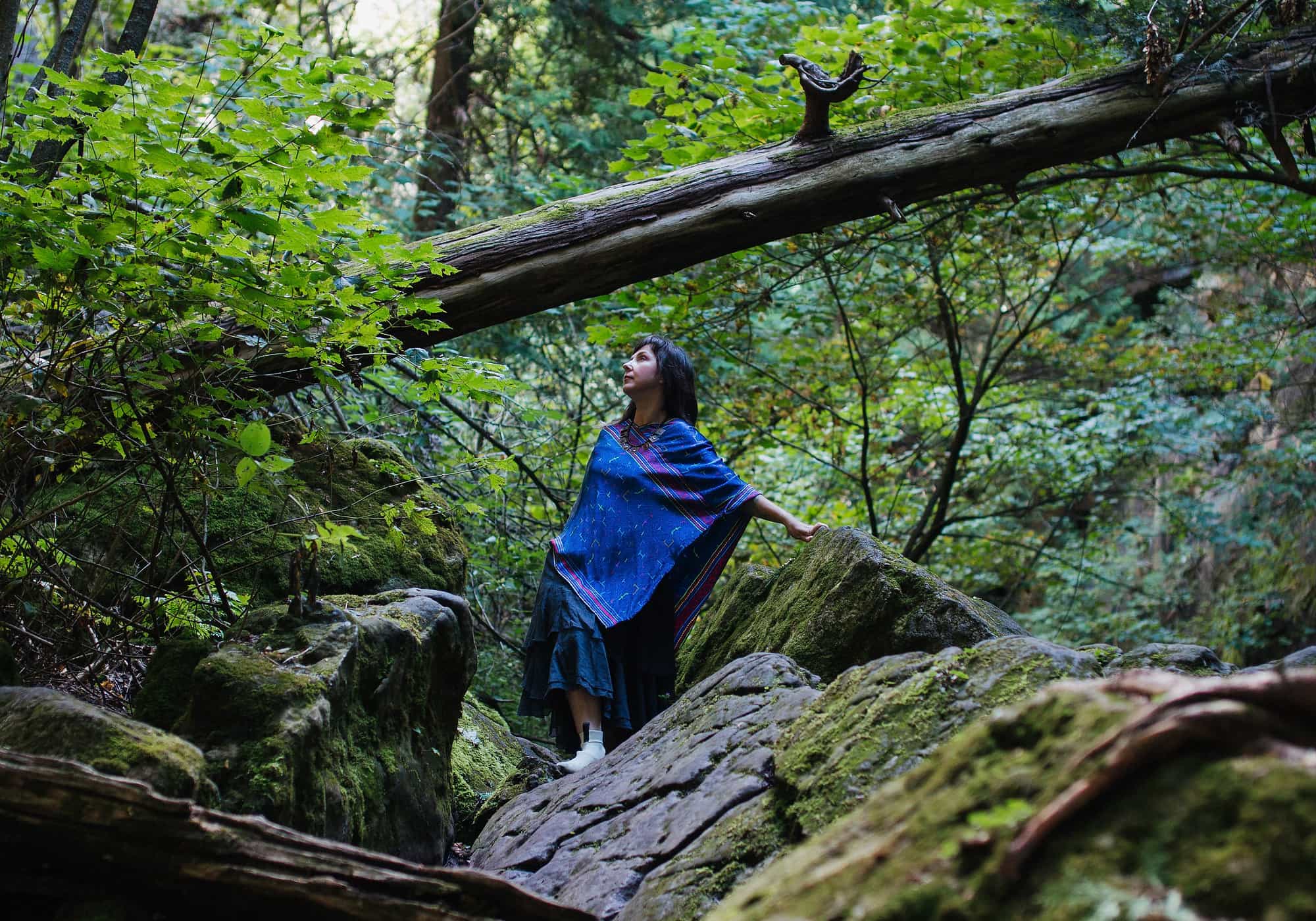 Mariya Garnet in a cobalt shawl standing among mossy boulders in old forest