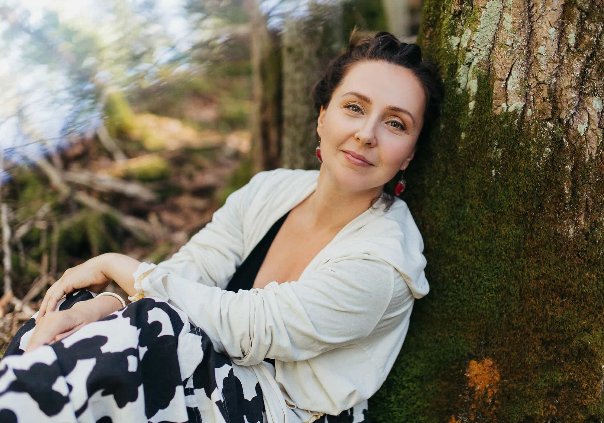 Mariya Garnet sitting against a tree trunk, smiling, in a sunlit forest